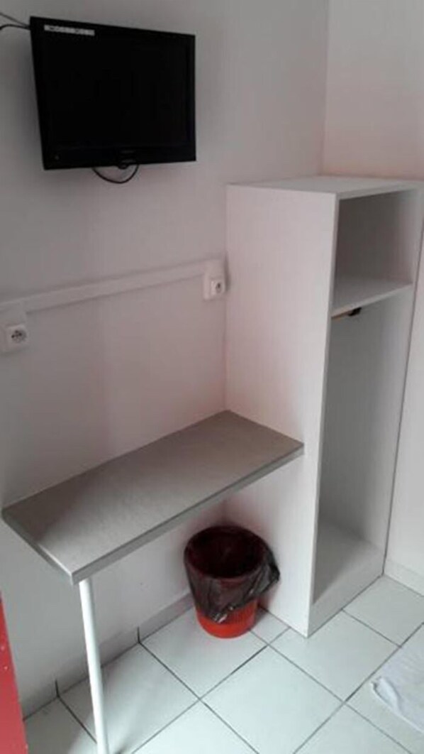 A small, empty room with a white shelf, a red bucket, and a black television mounted on the wall. - Hotel Du Centre (Saint-Denis)