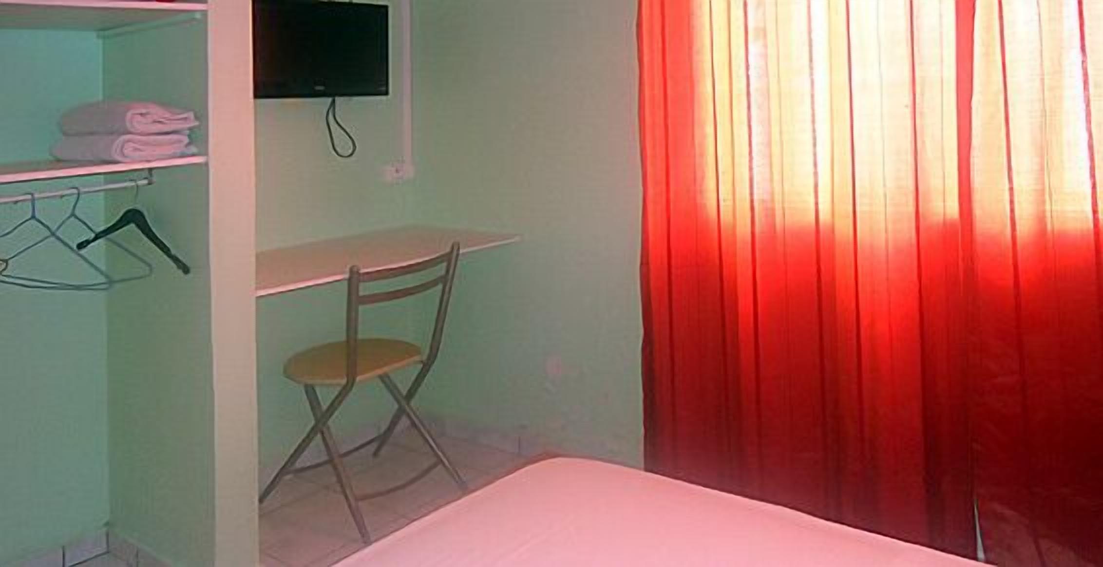 Una habitación con cama, escritorio, silla, televisor montado en la pared y una cortina roja.