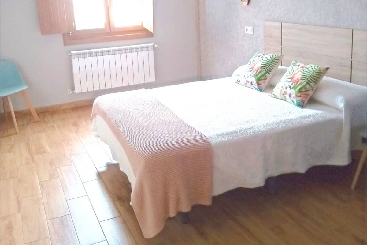 Un dormitorio con cama, ventana con cortinas, radiador y una silla.