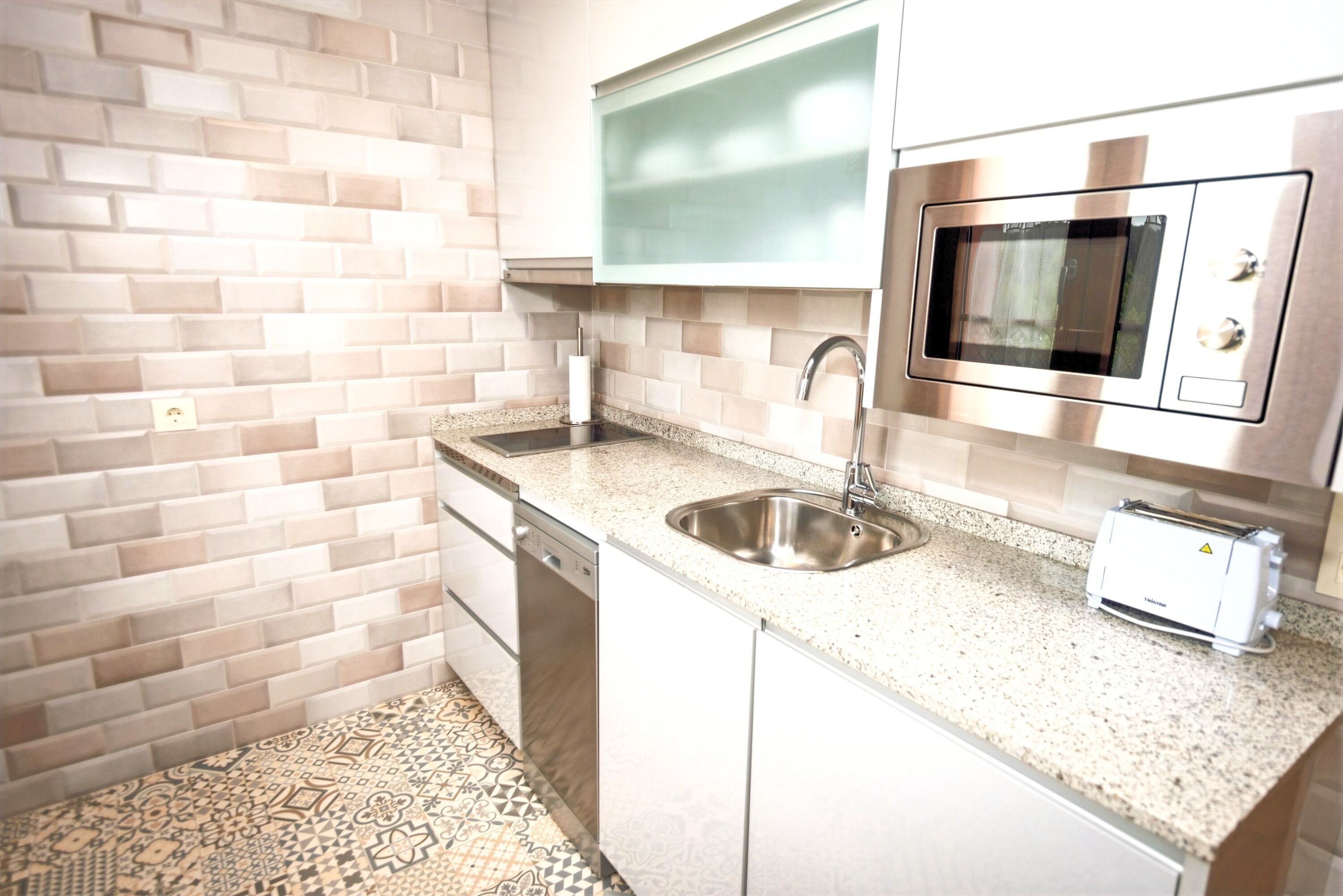 Una cocina compacta con un lavamanos de acero inoxidable, un microondas y un tostador. El salpicadero está hecho con baldosas beige, y el piso tiene un diseño de baldosas estampado.