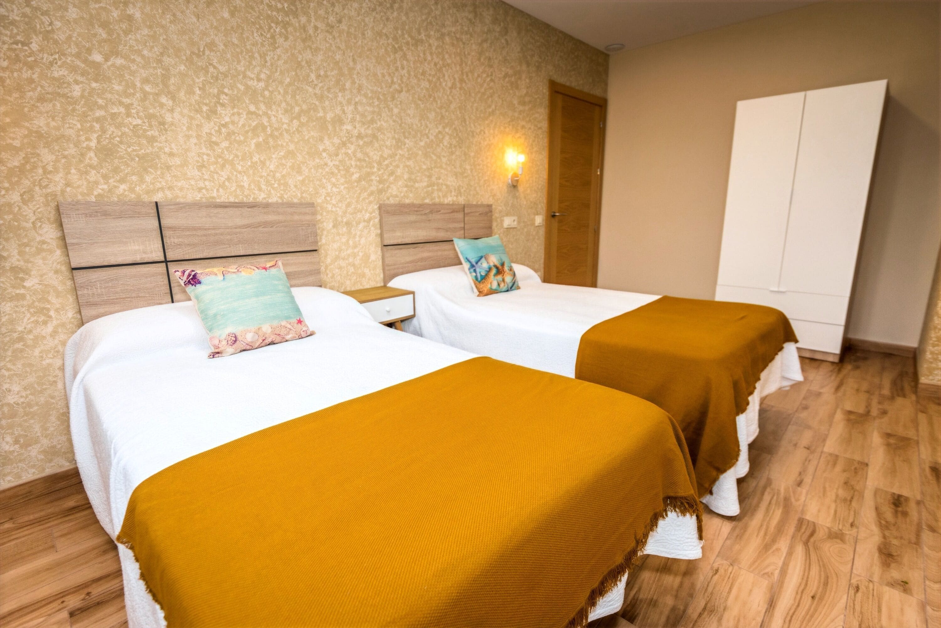 Una habitación de hotel con dos camas, cada una con cubrecama blanco y manta color mostaza. Tiene cabecera de madera, mesita de noche con lámpara y armario.
