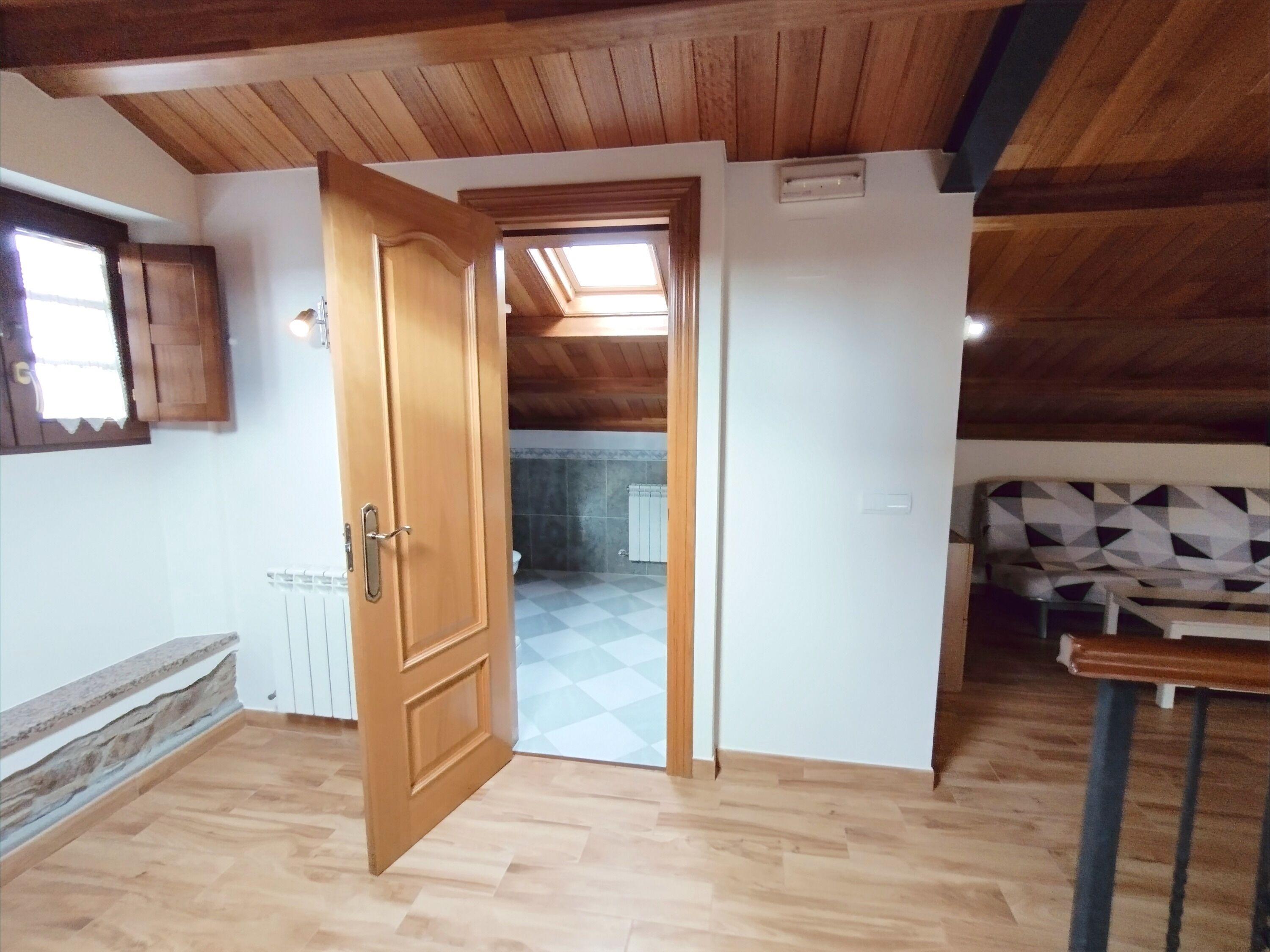 Una habitación con vigas de madera en el techo, una puerta de madera, suelo de cuadros y vistas a otra habitación con radiador a través de una puerta abierta.