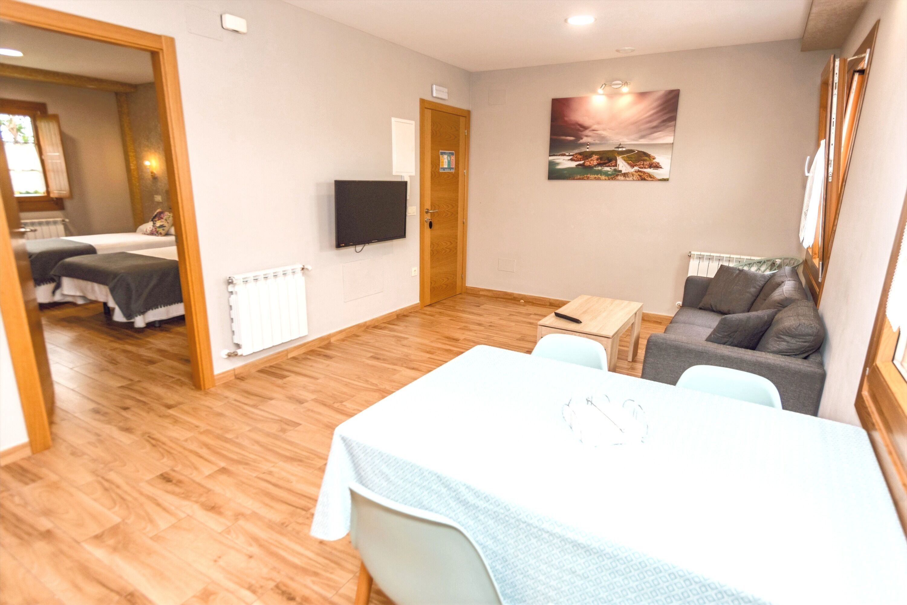 A hotel room with a wooden floor, a bed with a white headboard, a gray sofa, a small table, and a painting on the wall.