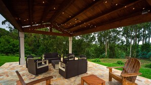 Un patio al aire libre con suelo de piedra, mobiliario de madera y luces colgantes.