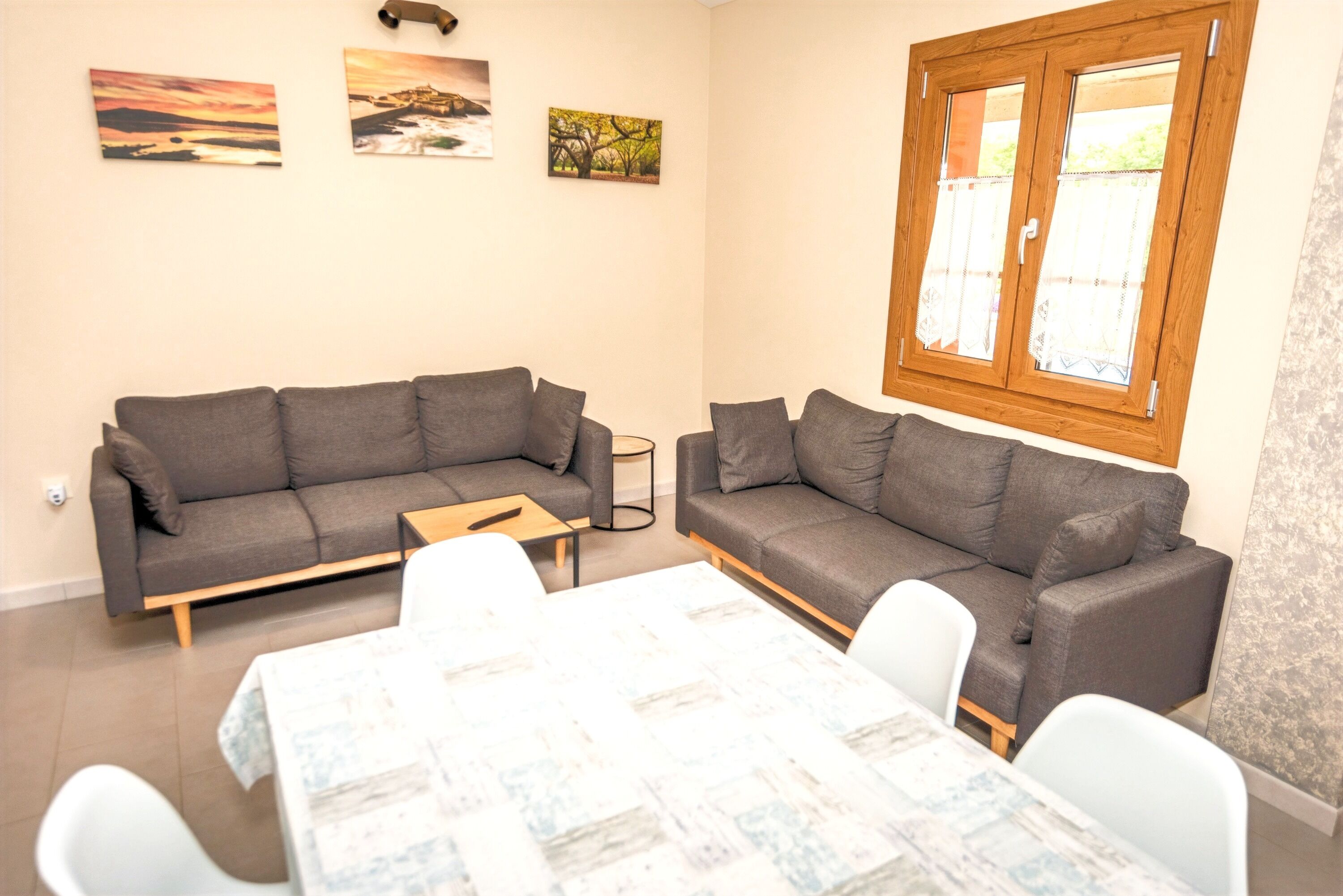 A living room with two grey sofas, a small table, and a dining table with chairs. There are three paintings on the wall and a window with wooden frames.
