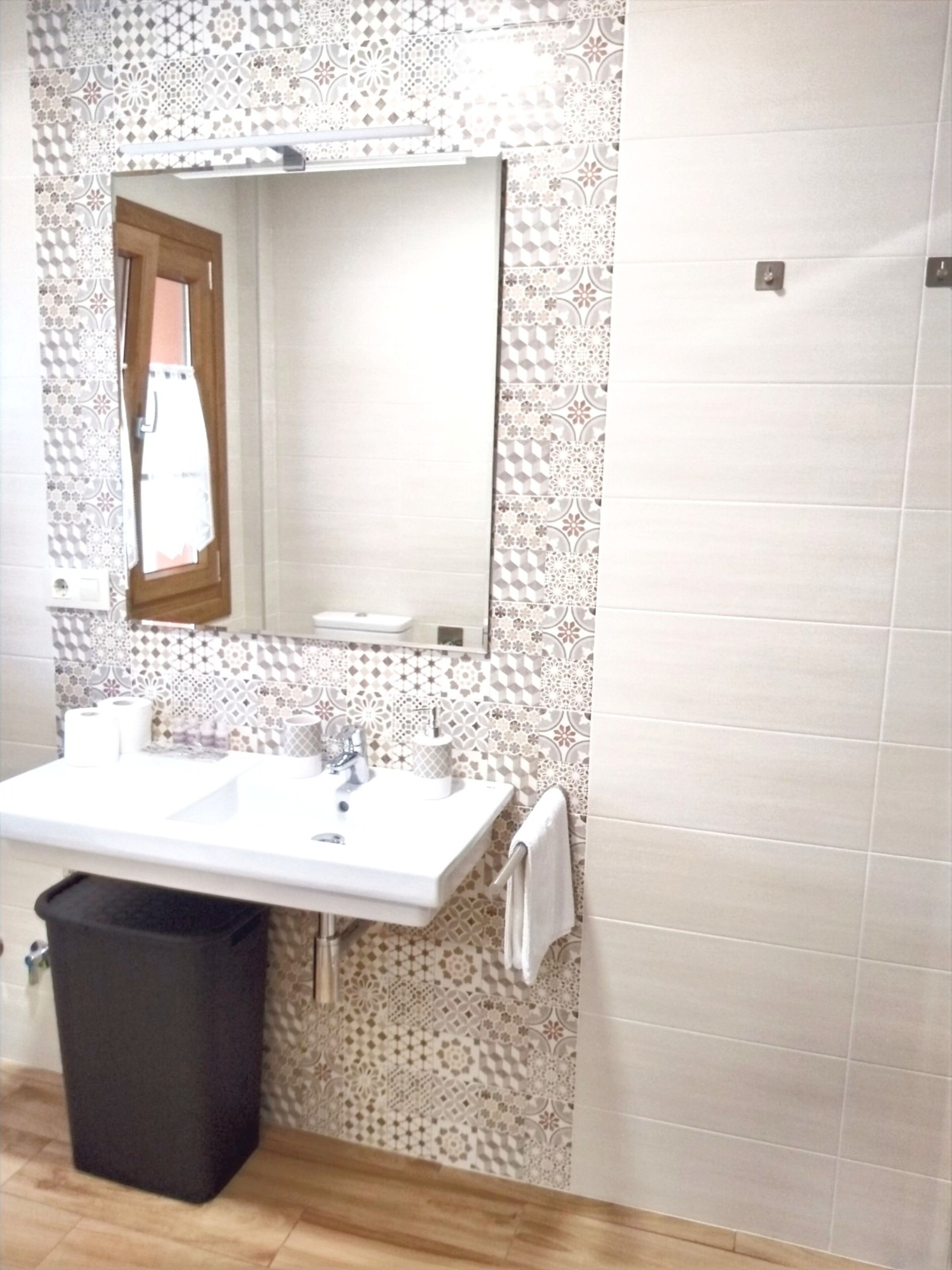 A bathroom with a white sink, a mirror, patterned tiles, and a black trash bin.