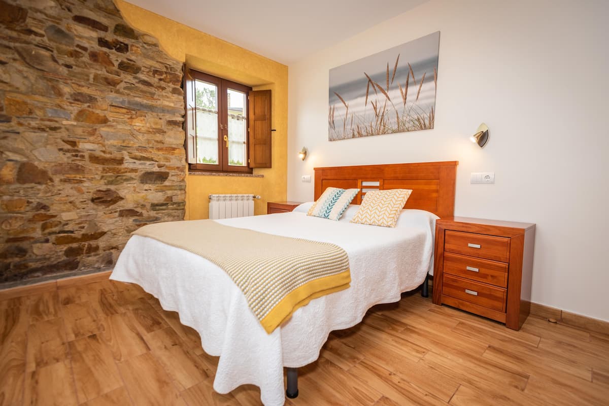 Un dormitorio con pared de piedra, suelo de madera, una cama con cabecero de madera, una mesita de noche y un cuadro en la pared.