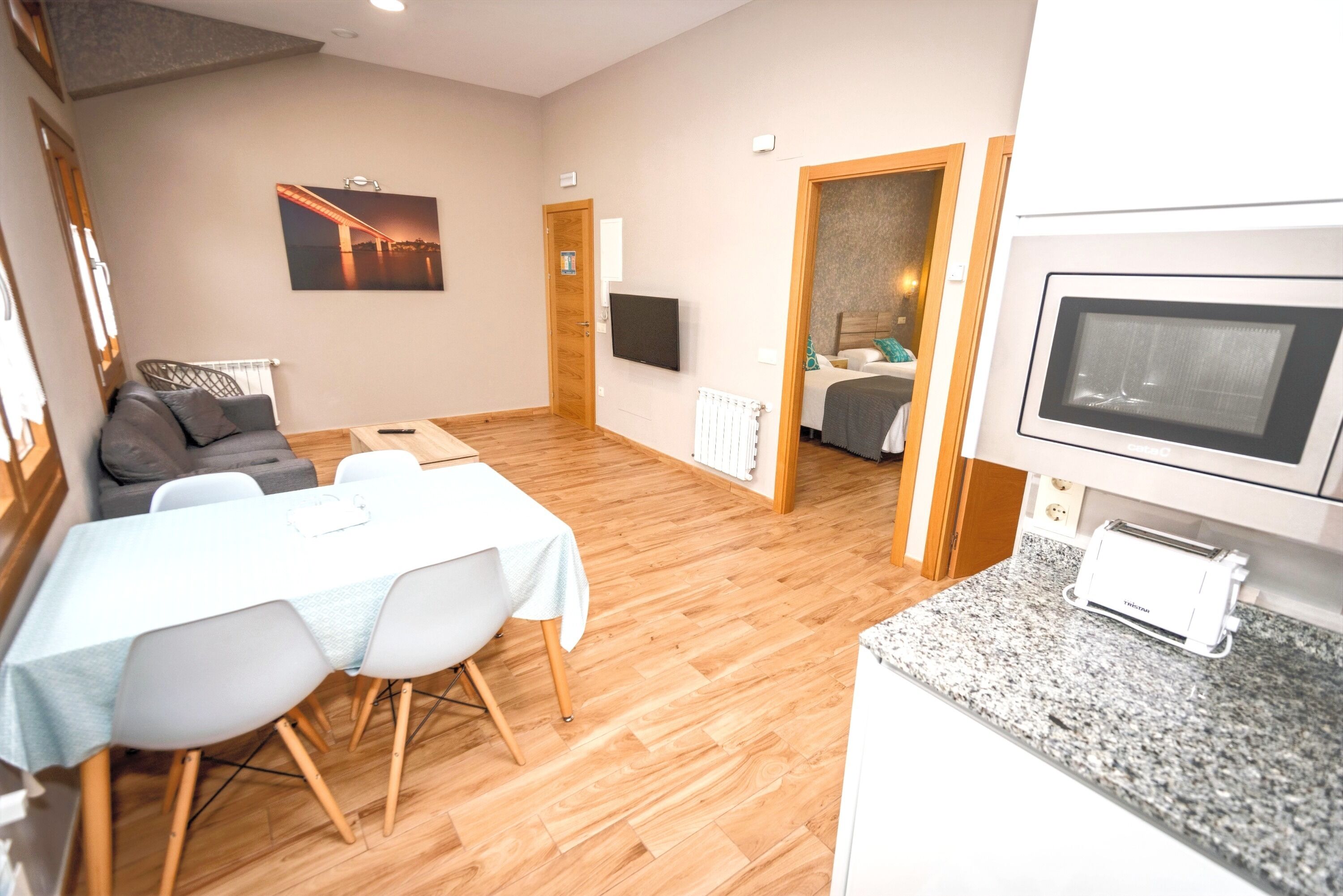 Una sala moderna con sofá, mesa de comedor y sillas. Hay un televisor montado en la pared, una zona de cocina con microondas y una puerta que da a otra habitación.