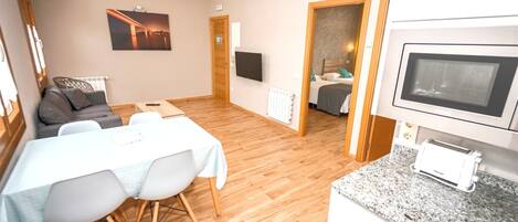 Una sala moderna con sofá, mesa de comedor y sillas. Hay un televisor montado en la pared, una zona de cocina con microondas y una puerta que da a otra habitación.