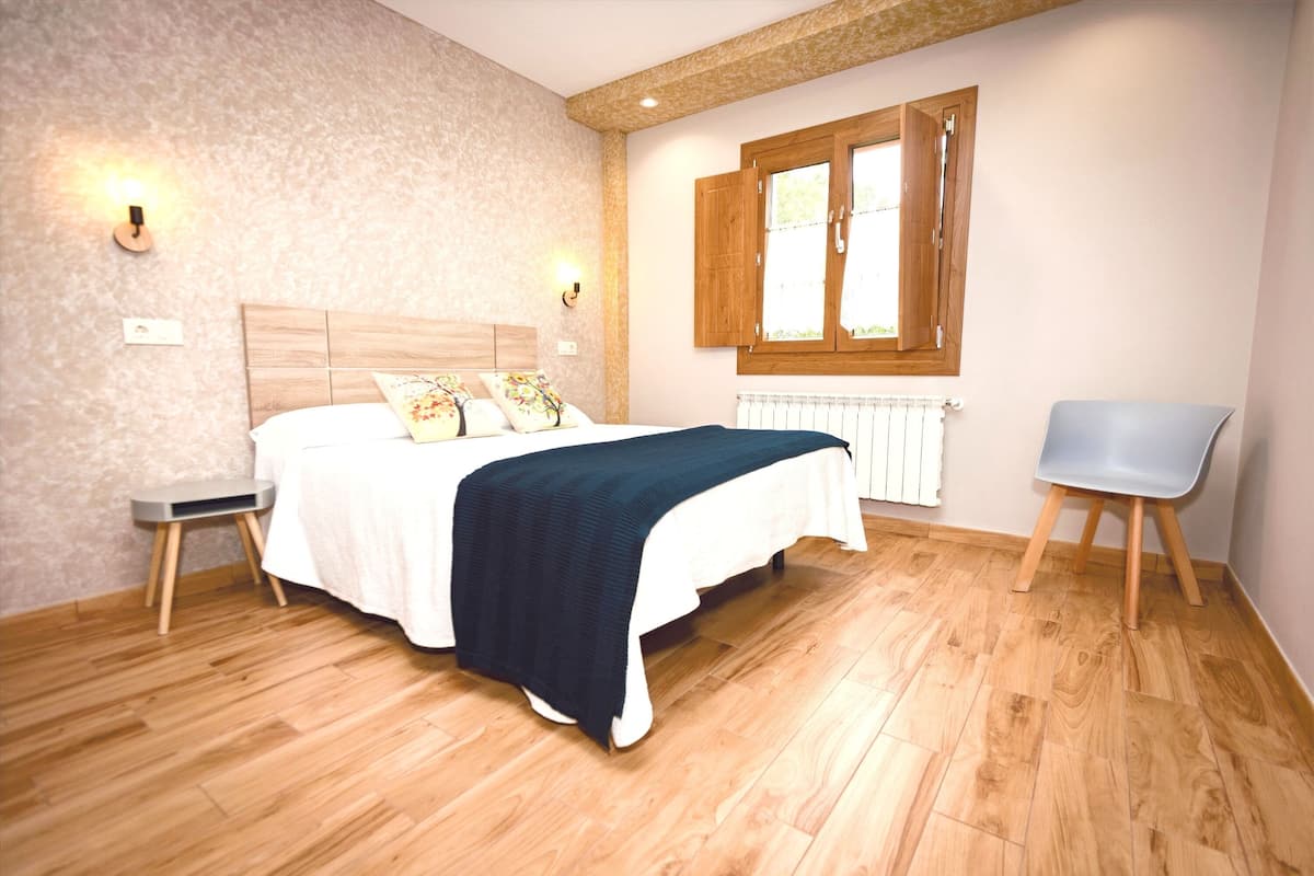 Un dormitorio con una cama, mesitas de noche, una silla, una ventana con contraventanas de madera y un radiador.