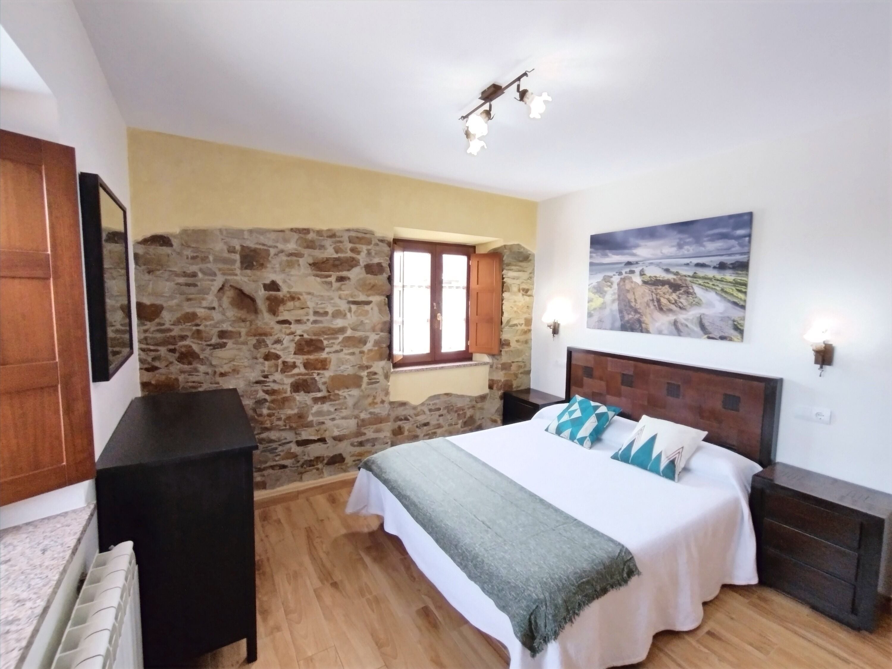 Een slaapkamer met een stenen muur, een bed met een wit en grijs beddegoed, een houten hoofdbord, en een schilderij van een landschap.