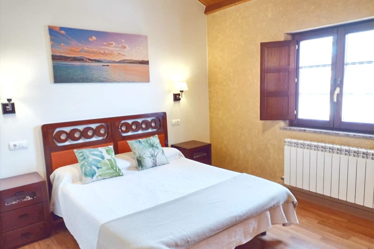 Un dormitorio con cama de madera, una mesita de noche, un cuadro colgado en la pared, una ventana con contraventanas de madera y un radiador.