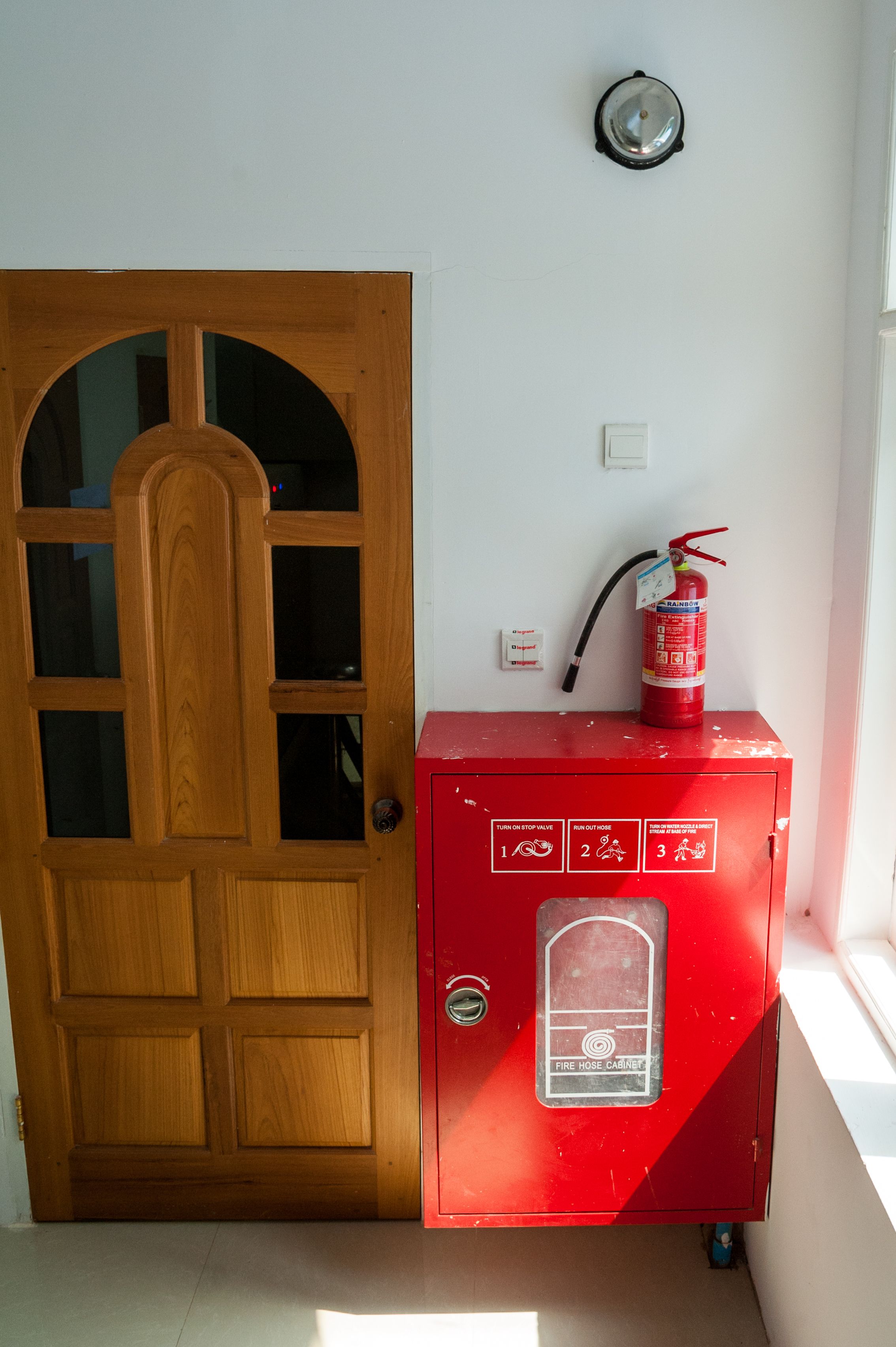 Une porte en bois avec un panneau de verre, un coffre rouge contenant un tuyau d’incendie et un extincteur, et un détecteur de fumée fixé au mur.