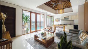 A spacious living area with a large sliding glass door leading to a pool, a dining area with wooden furniture, a kitchen with white cabinets, and a ceiling fan. - Kakul Villas & Suites (Ubud)
