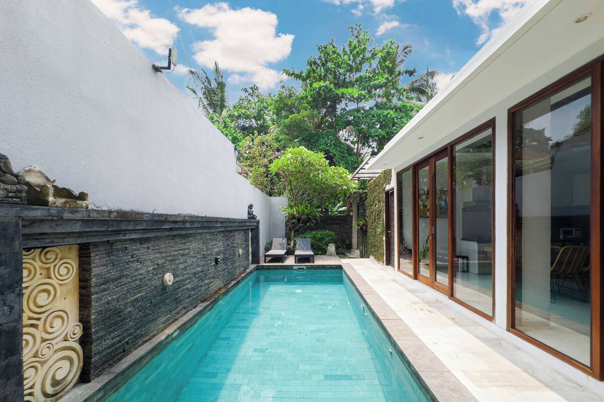 A rectangular swimming pool with clear blue water, surrounded by a stone wall with decorative patterns, a modern house with large glass doors, and a lush green garden with trees and plants.