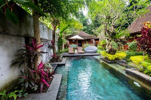 A serene pool area with a small house, surrounded by lush greenery and a clear pool.