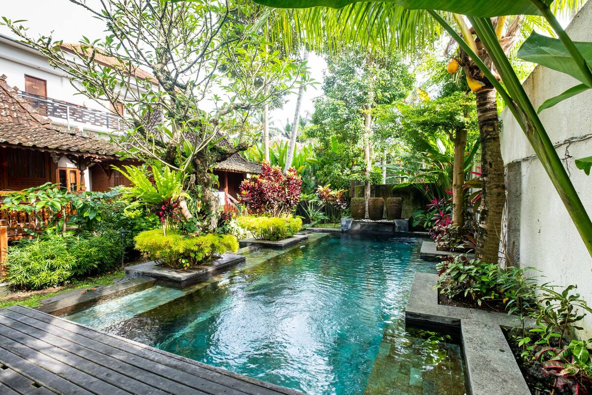 A serene pool area with a wooden deck, surrounded by lush greenery and a small traditional hut.