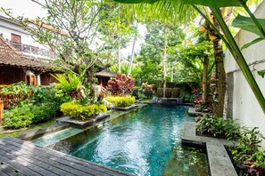 A serene pool area with a wooden deck, surrounded by lush greenery and a small traditional hut.