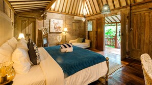 A cozy bedroom with a wooden ceiling, a bed with white bedding and a blue blanket, a bedside table with a lamp, and a view of the outdoors through a door. - Kakul Villas & Suites (Ubud)