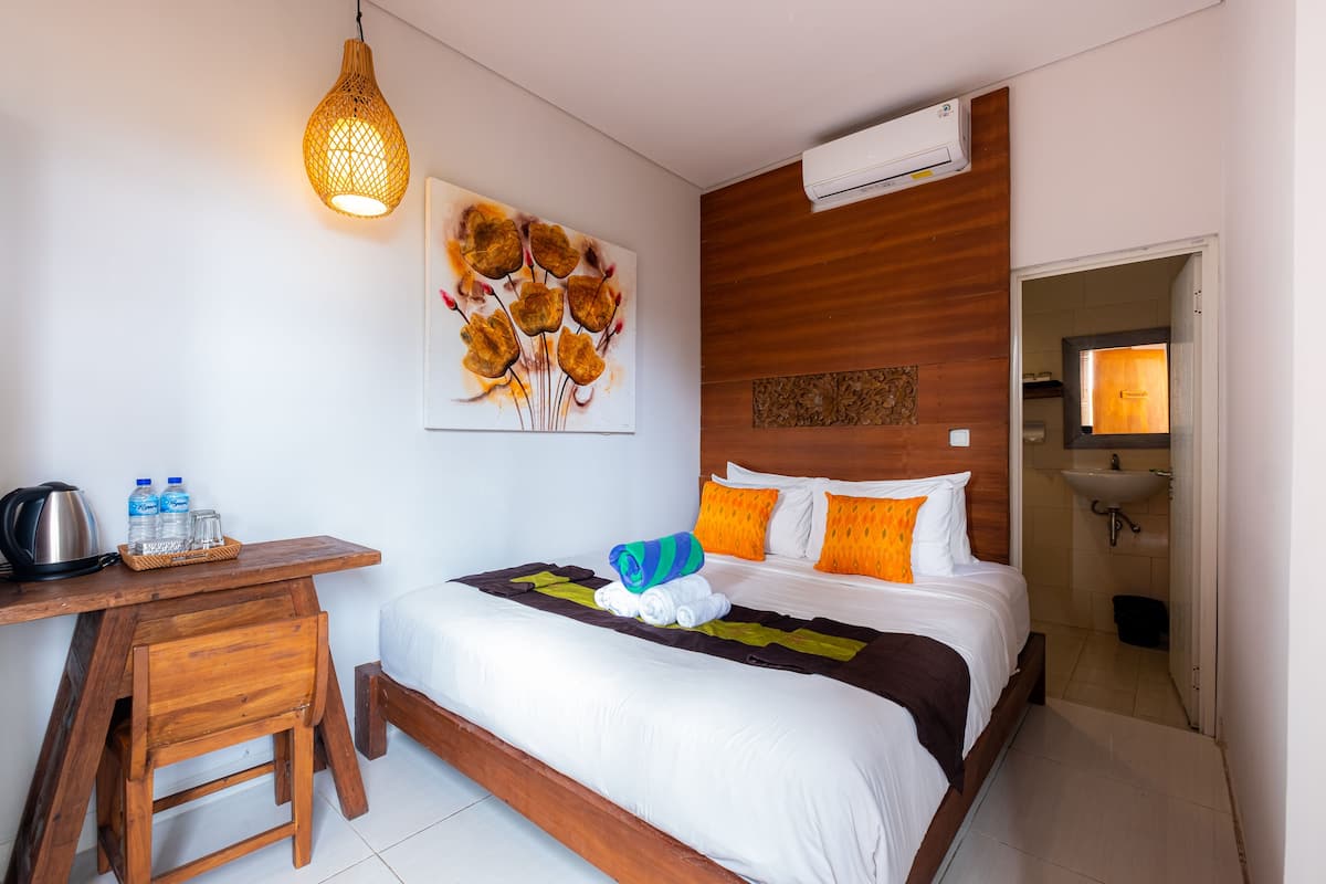 A hotel room with a wooden desk, a bed with white and orange bedding, a painting on the wall, and an air conditioning unit.