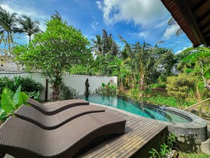 A pool with a wooden deck and lounge chairs, surrounded by lush greenery and a clear sky.
