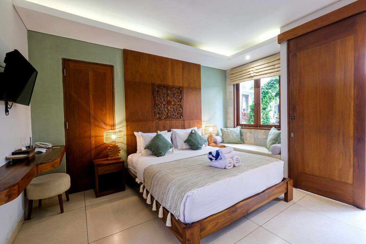 A bedroom with a wooden bed, bedside tables, a TV, and a window with a view of greenery.
