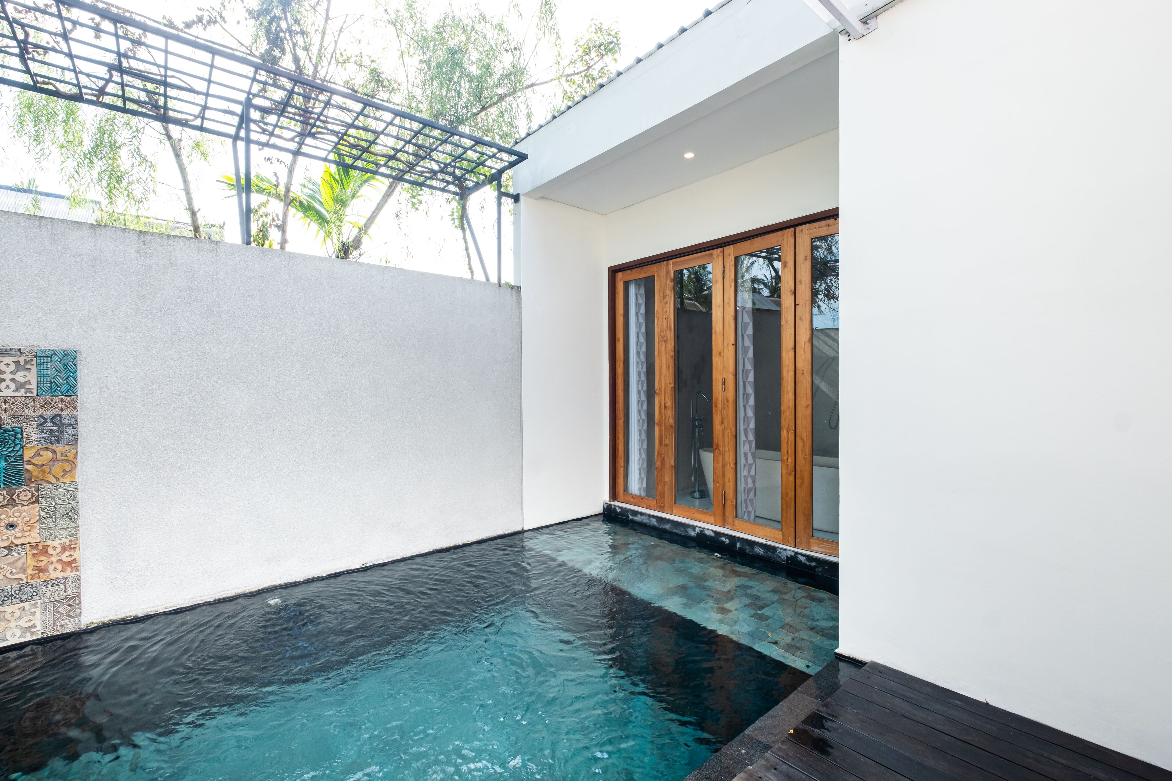 A modern outdoor swimming pool area with a wooden deck, a glass door entrance, and a pergola structure.