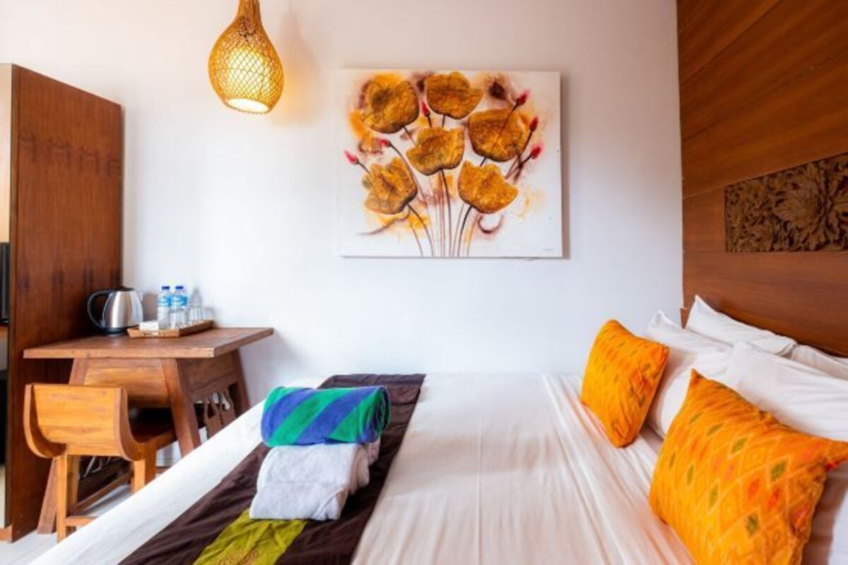 A hotel room with a bed, a desk, a chair, a painting on the wall, and a hanging light fixture.