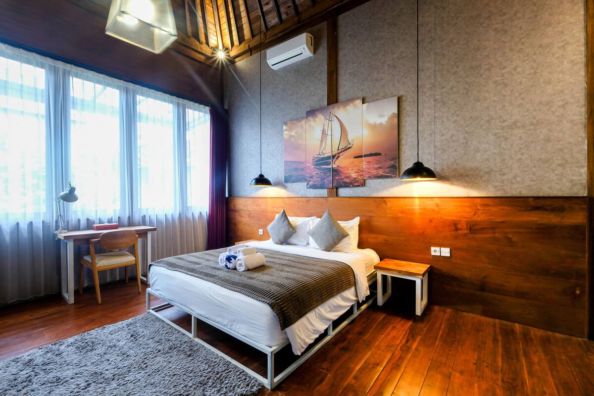 A bedroom with a wooden floor, a bed with white linens, a desk, and a chair.
