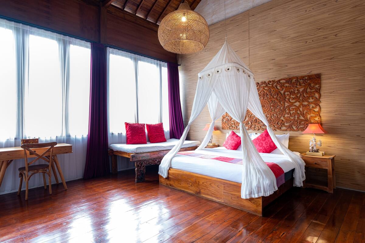 A wooden-floored bedroom with a four-poster bed, a wooden desk, and a large window with curtains.