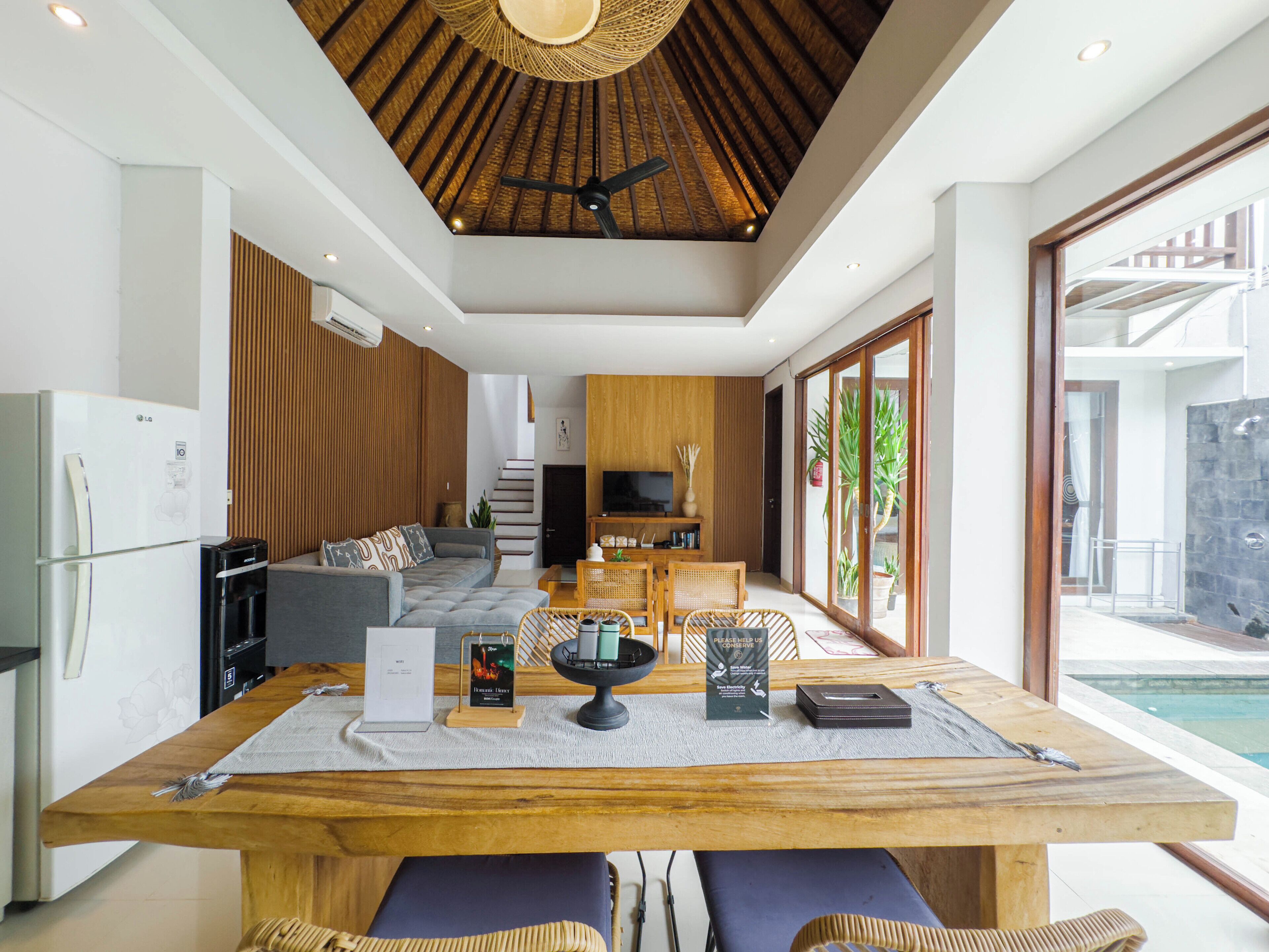 A modern living room with a wooden ceiling, a dining area with a table set for two, and a view of the pool outside.
