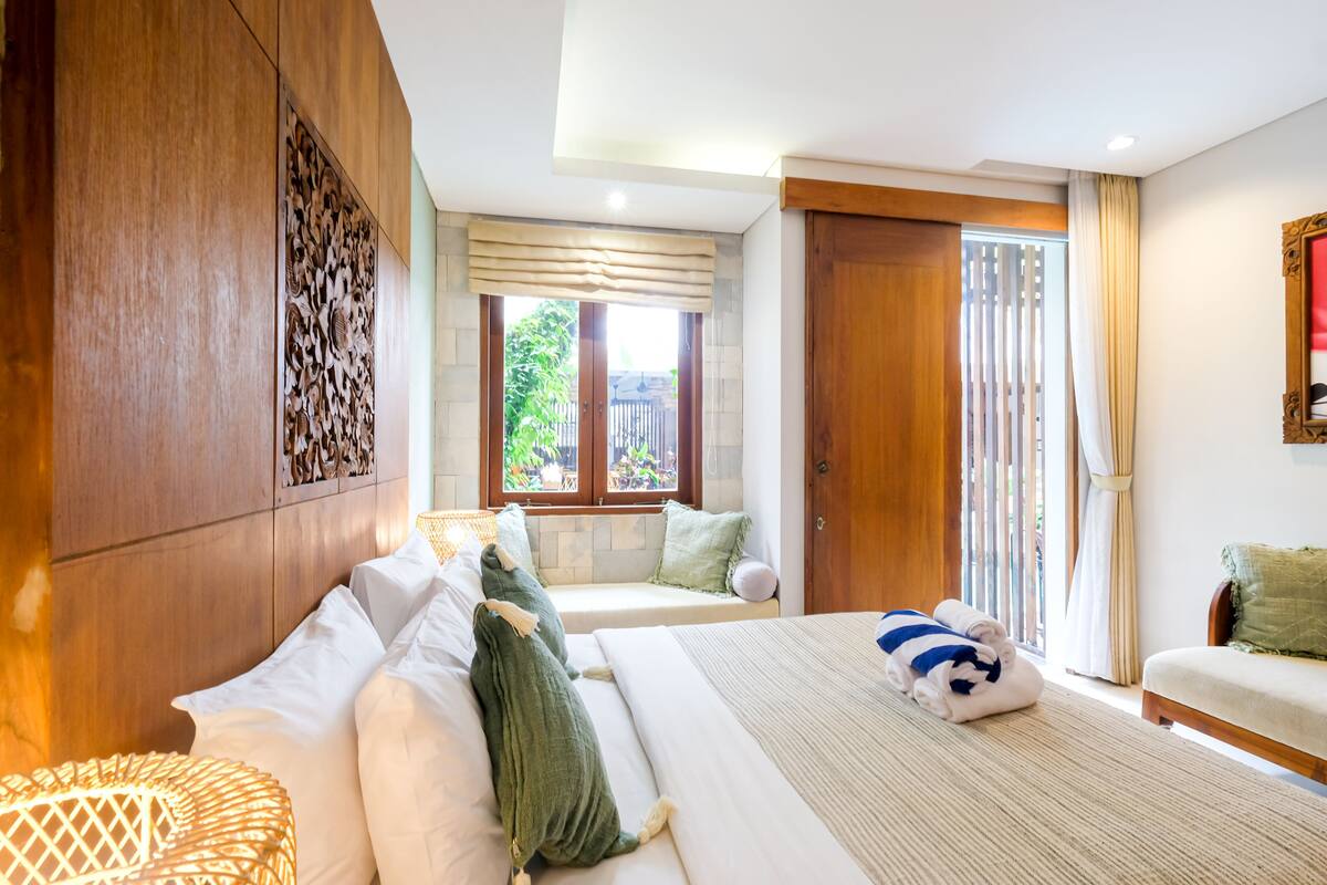 A hotel room with a large bed, a wooden headboard, a window with a view of greenery, and a sliding door.