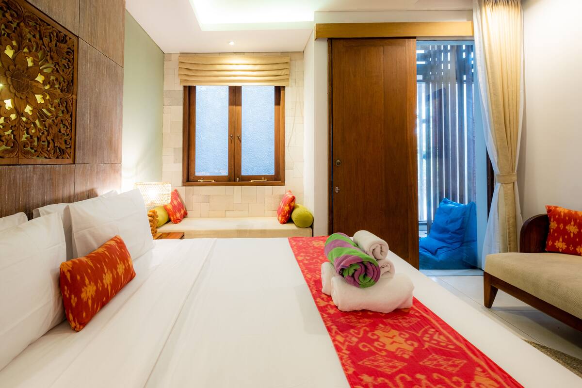 A hotel room with a large bed, a wooden headboard, a red and white patterned area rug, a wooden door, and a view of the outside through a window with curtains.