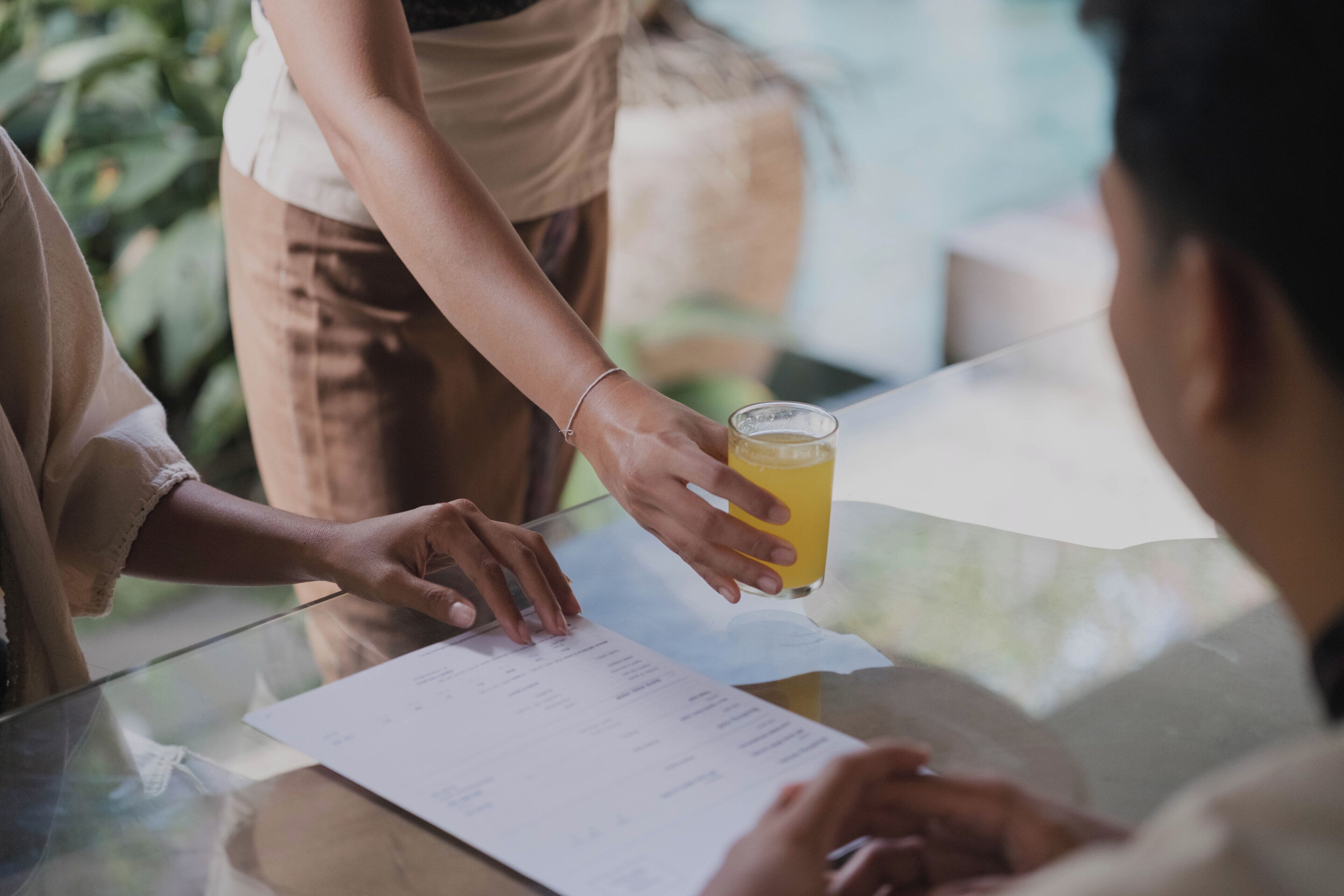 A person holding a glass of orange juice, a menu, and a tablet on a glass table.