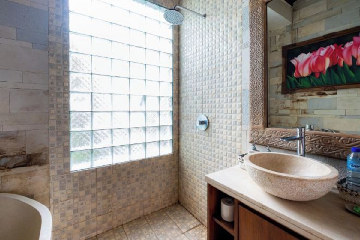 A bathroom with a large window, a bathtub, a sink with a stone basin, and a mirror with a floral painting.
