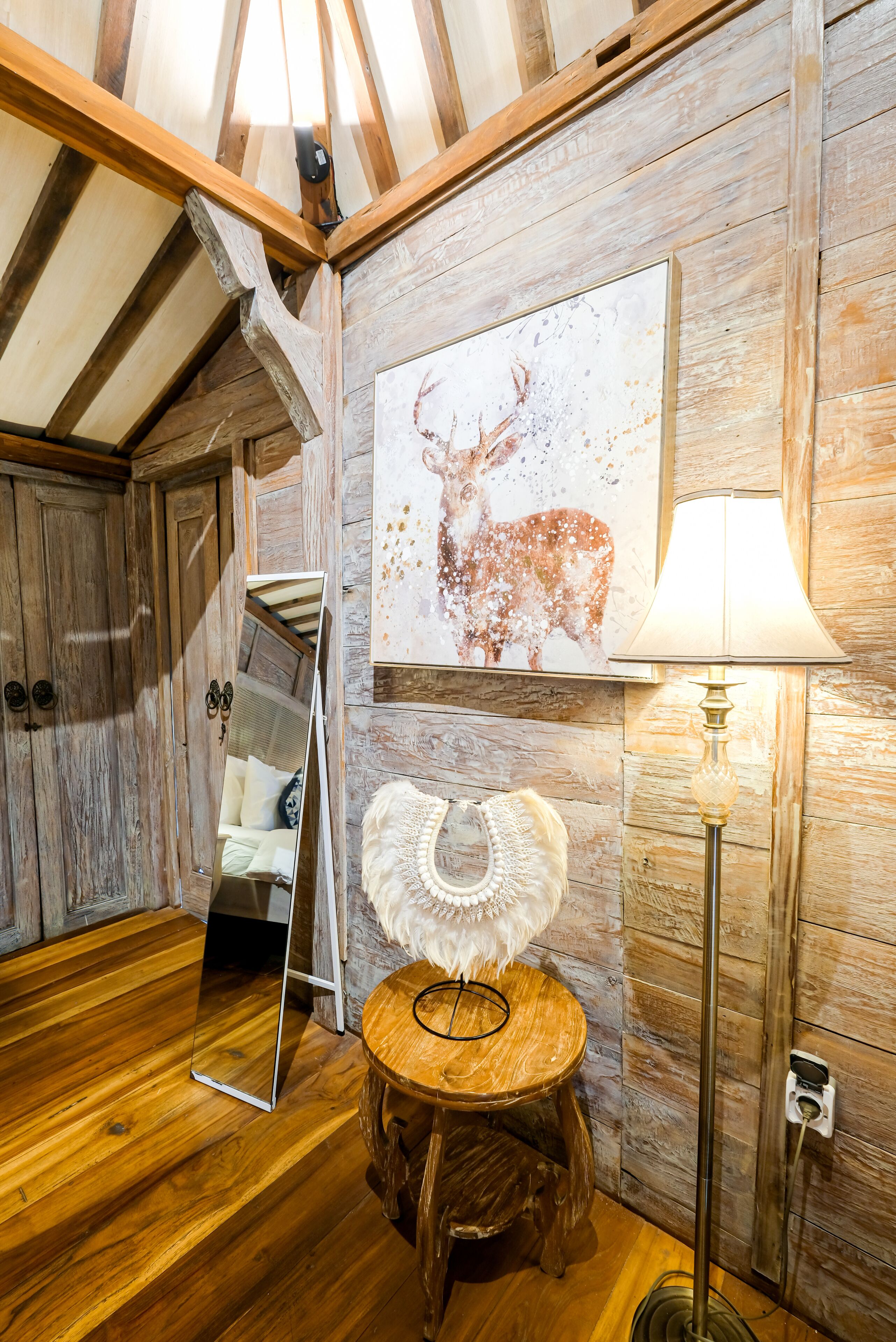 A rustic room with wooden walls, a wooden ceiling, a mirror, a lamp, and a painting of a deer.