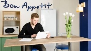 Une personne est assise à une table devant un ordinateur portable, des papiers et un vase de fleurs. On aperçoit un four à micro-ondes sur la gauche et un réfrigérateur sur la droite. Sur le mur derrière, on peut lire une inscription : « Bon appétit ».