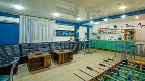 Une pièce comprenant un canapé en cuir noir, une table basse en bois, un baby-foot et un coin cuisine doté d’un plan de travail vert et de tabourets de bar.
