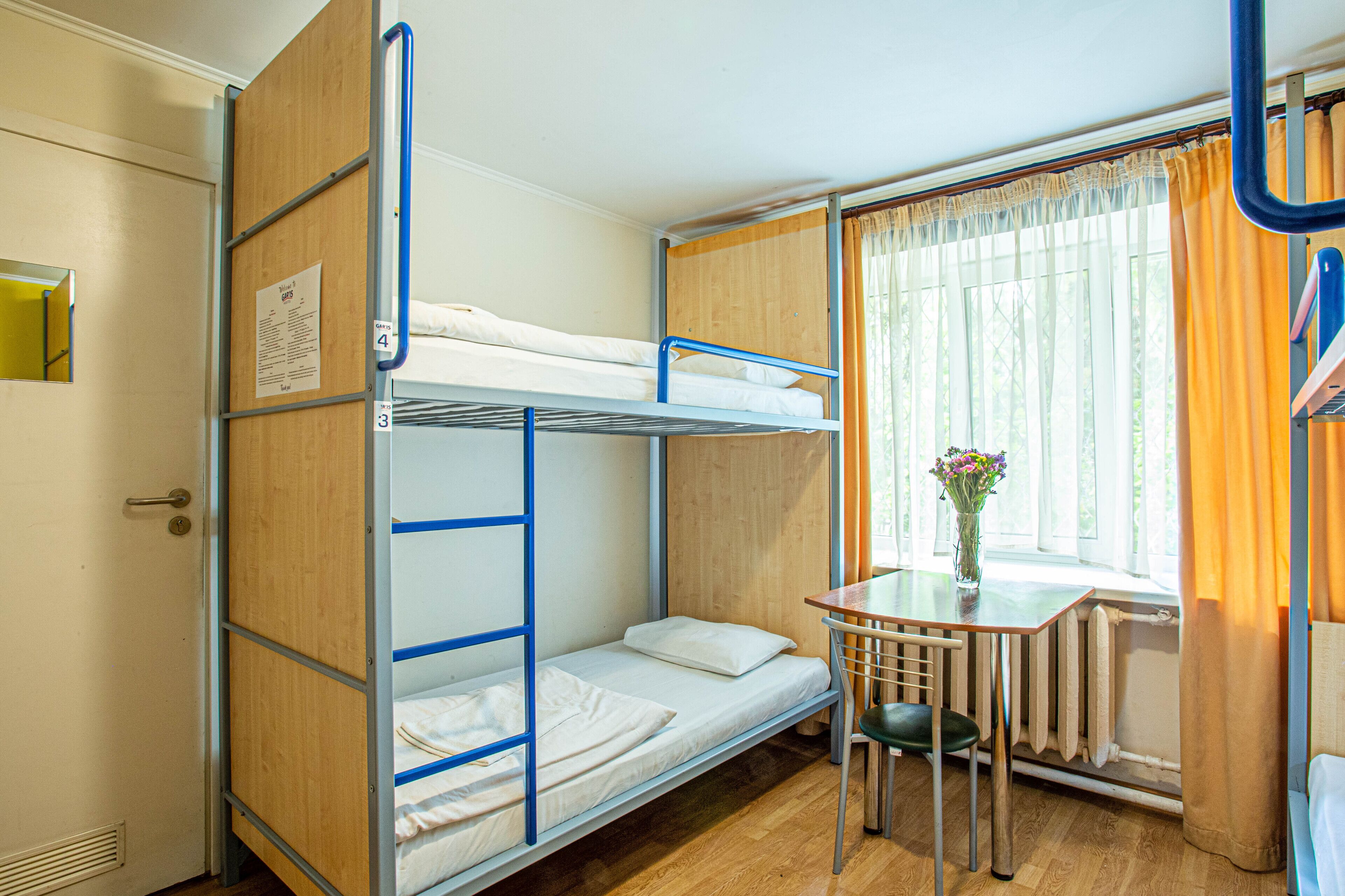 A bunk bed room with a small table and chair, a window with curtains, and a radiator.