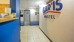 Un couloir avec un comptoir d’accueil, une pancarte indiquant « Bienvenue à l’auberge GARIS », et une porte donnant accès aux chambres.