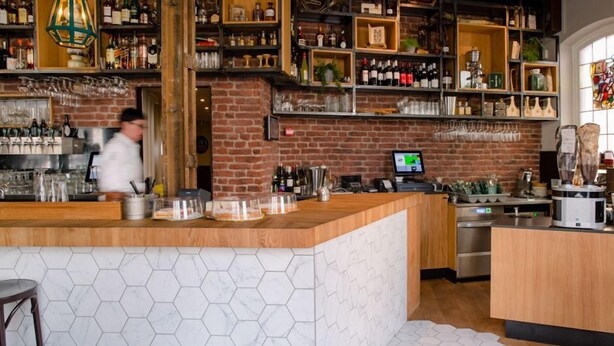 Un espace bar avec un mur en briques, des étagères remplies de bouteilles, une caisse enregistreuse et une personne à l’arrière-plan.