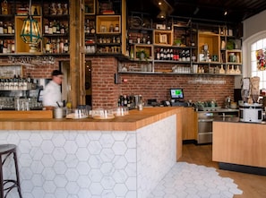 Eine Bar mit Ziegelwand, Regalen voller Flaschen, einer Kasse und einer Person im Hintergrund.