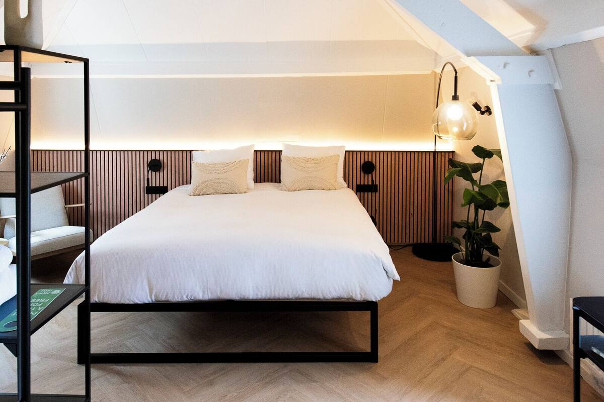 A modern loft bedroom with a sloped ceiling, a large bed, a bedside table, a lamp, and a potted plant.
