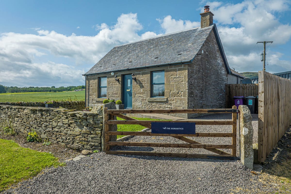 A stone cottage with a wooden gate, a stone wall, and a gravel driveway.