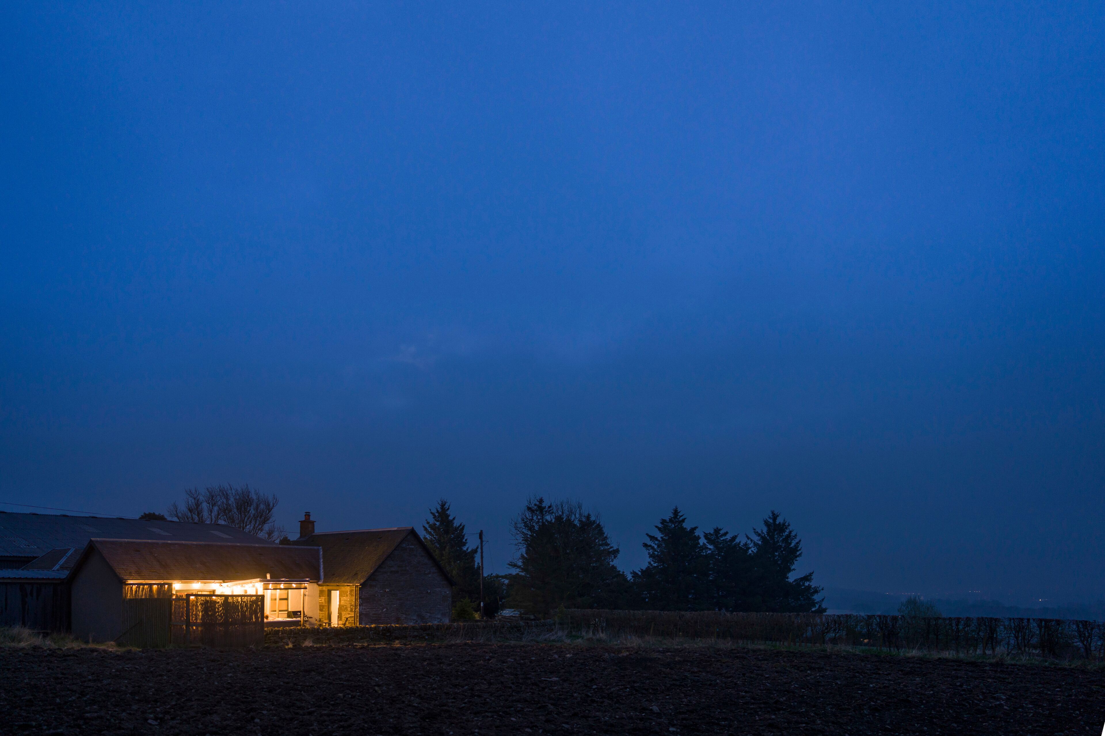 A rural house with a lit interior, surrounded by fields and trees.