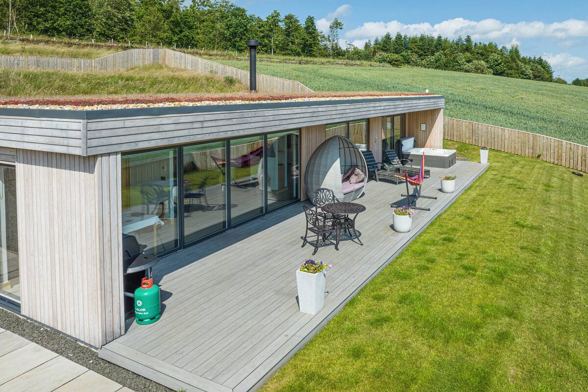 A modern house with a large deck, outdoor seating, and a view of a grassy field.