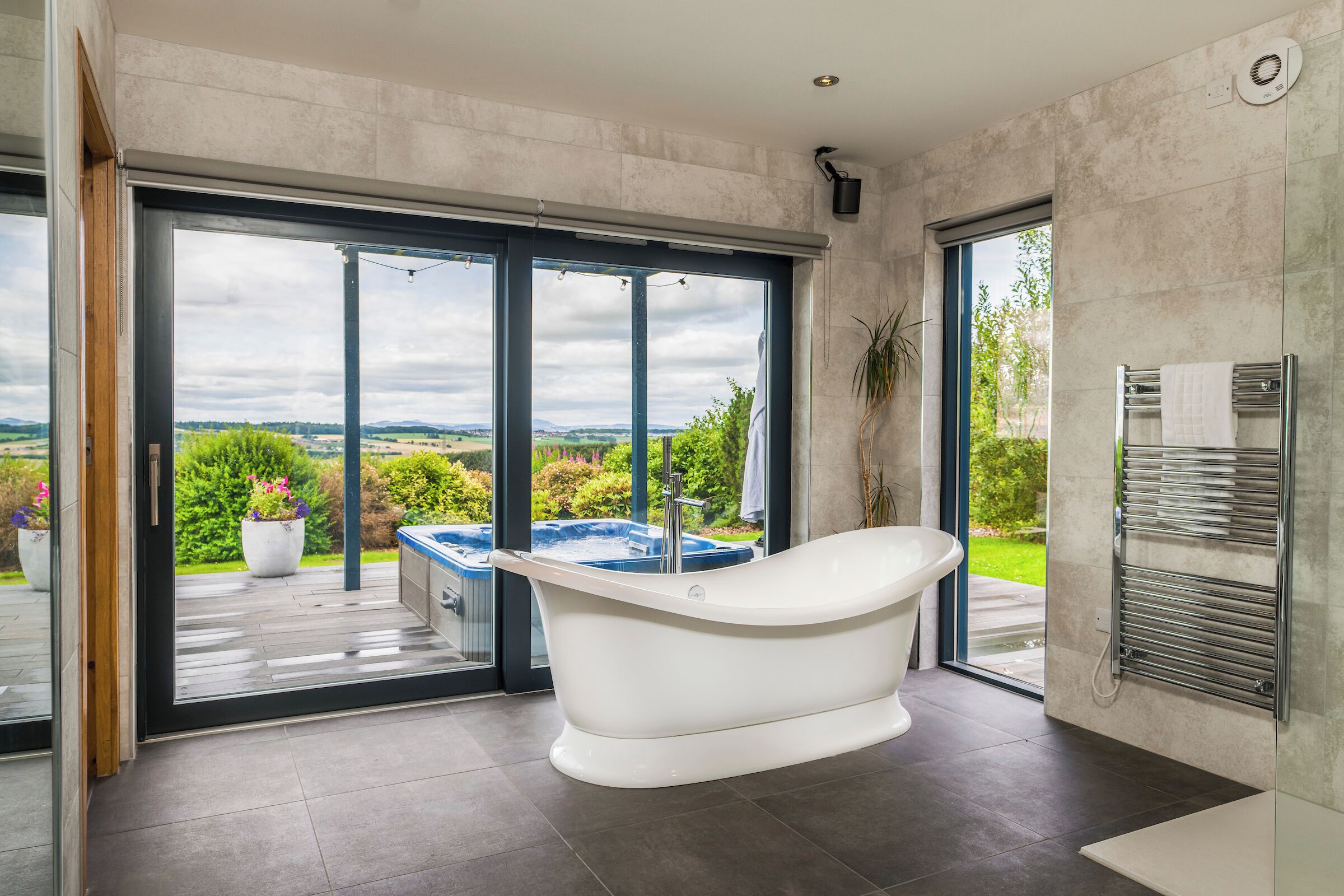 A modern bathroom with a freestanding bathtub, large sliding glass doors leading to a patio with a hot tub, and a view of the countryside.