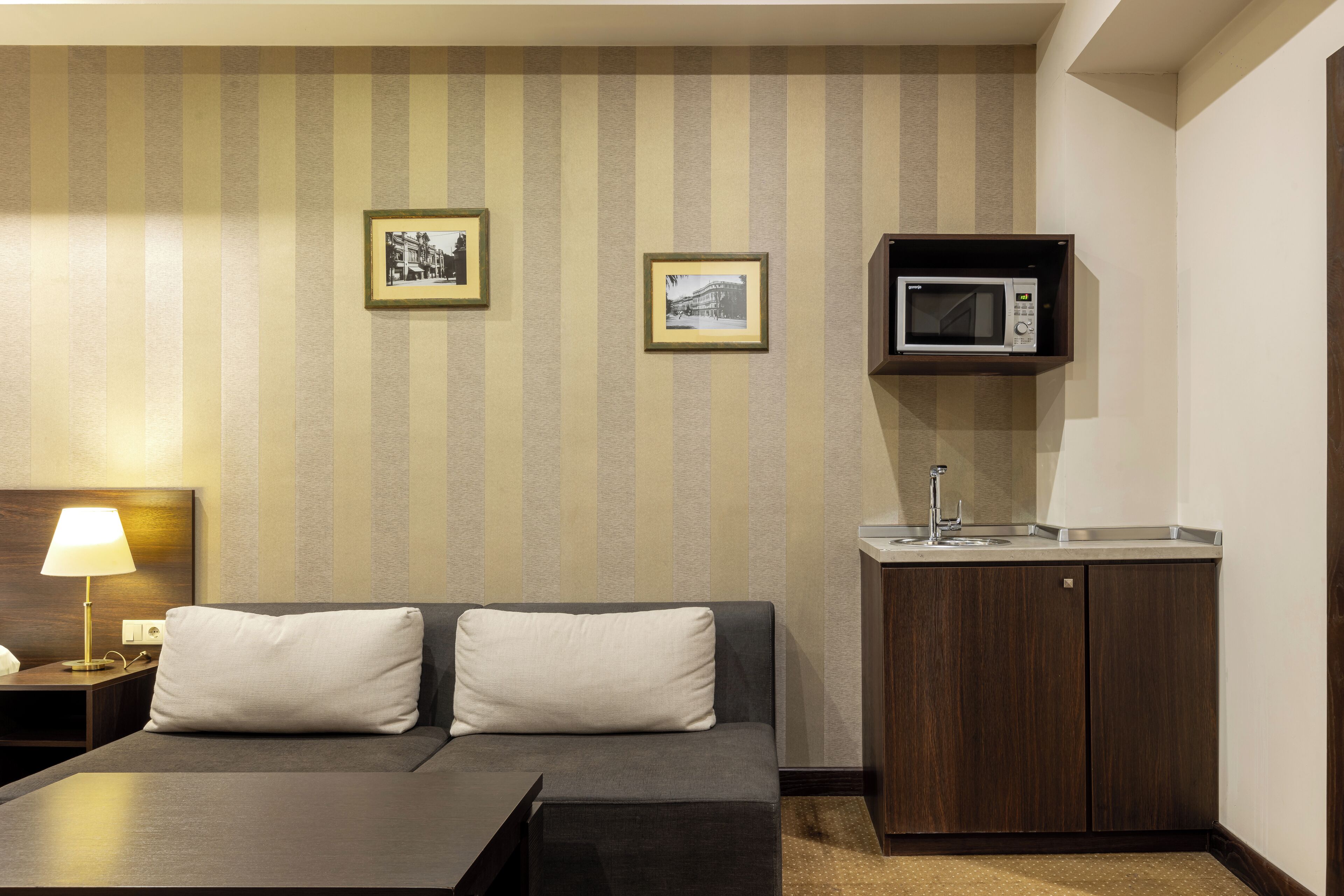 A hotel room with a sofa, a small kitchenette, and two framed pictures on the wall.
