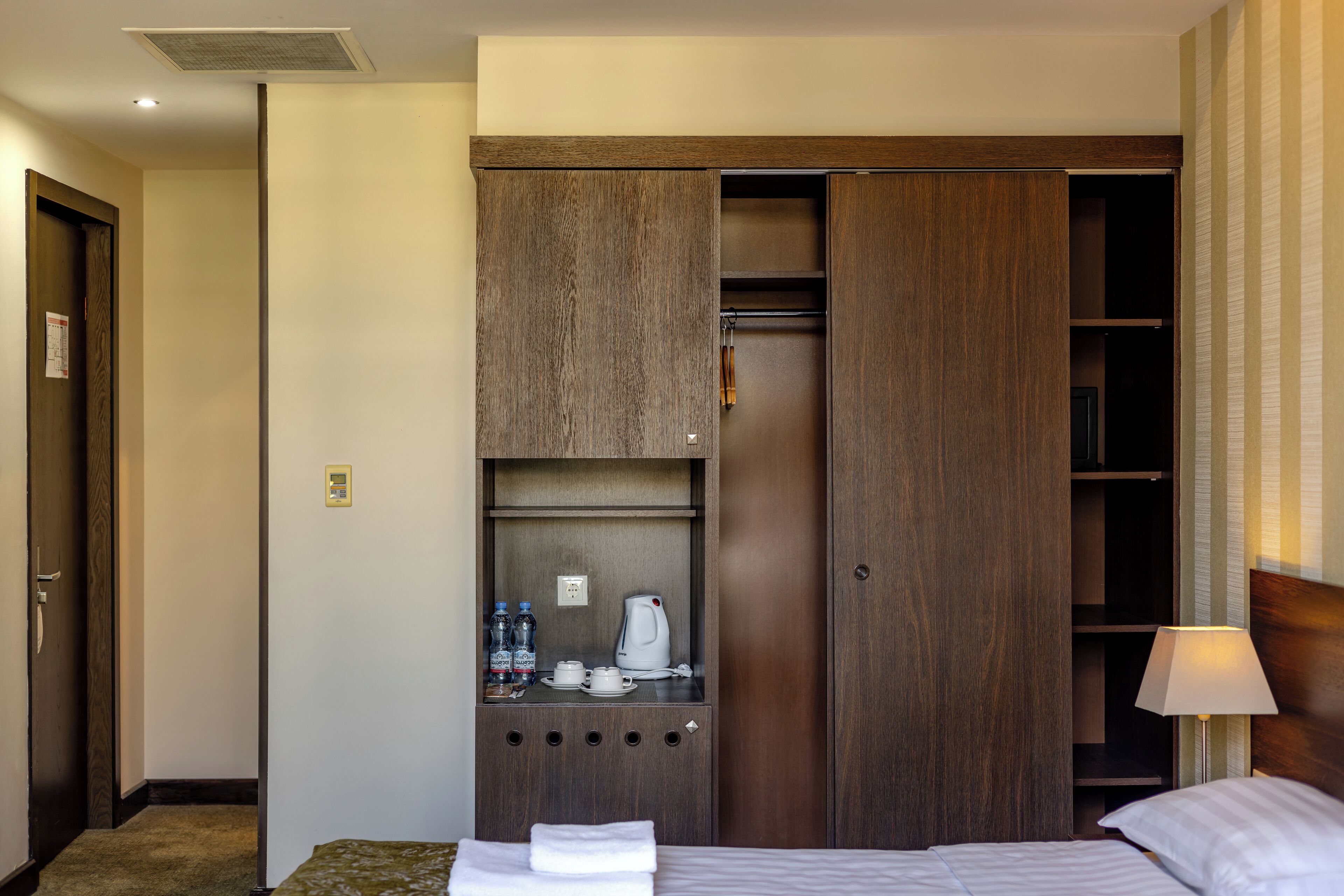 A hotel room with a bed, a wardrobe with a mirror, a small table with a kettle, and a nightstand with a lamp.