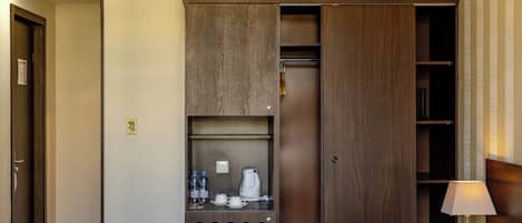 Quarto de hotel com cama, guarda-roupa com espelho, mesa pequena com chaleira e mesa de cabeceira com abajur.